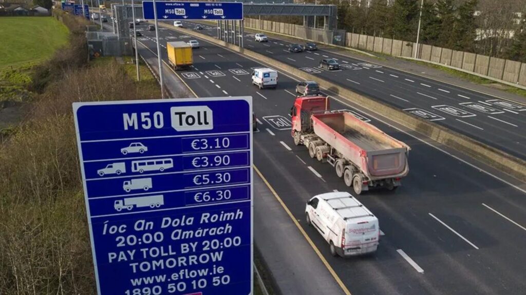 The image shows the M50 motorway with many cars on the road. There is a sign showing all the costs of the M50 toll for each size car, along with a notice to pay by 8 p.m. the next day.