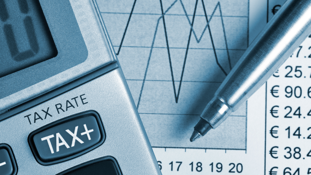 A close-up image showing a part of a calculator with buttons labelled 'TAX RATE' and 'TAX+' in focus, alongside a pen lying on top of a printed financial spreadsheet with various numbers in Euros, indicating calculations related to taxes.